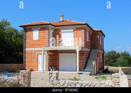 Unfinished briques rouges maison de famille avec de nouvelles portes et fenêtres entouré des murs en pierre et des matériaux de construction avec des arbres denses et ciel bleu clair Banque D'Images