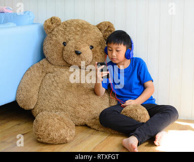 Petit garçon asiatique dans l'aide de casque smartphone avec teddy à la maison Banque D'Images