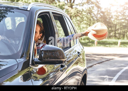 Coup de voiture brillante horizontale le long de la route avec un homme derrière une roue. Woman waving avec un chapeau dans sa main, alors que son mari est au volant d'une blac Banque D'Images