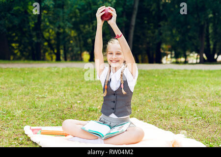 L'étudiant est souriant joyeusement et tient une pomme rouge au-dessus. lycéenne assis sur une couverture dans un parc avec des livres et à faire leurs devoirs Banque D'Images