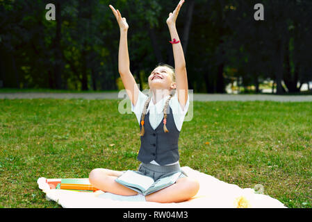 En étudiant sourit joyeusement uniforme et élevé au-dessus de sa tête à deux mains. écolière est assis sur une couverture dans un parc avec des livres et à faire leurs devoirs Banque D'Images