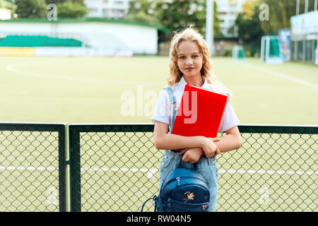 Lycéenne blonde aux yeux bleus tenant un dossier rouge et un sac à dos. se trouve près de l'étudiant sports stadium Banque D'Images