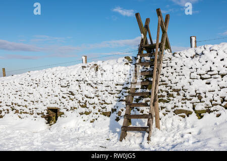 Mur de pierres sèches couvertes de neige dans le Derbyshire Peak District, l'échelle contre un ciel bleu froid. Banque D'Images
