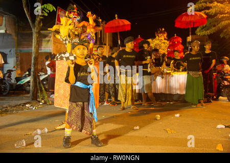 BALI, INDONÉSIE - Mars 27, 2017 : Garçon en costume traditionnel holding chiffres Nyepi, défilé traditionnel dans les rues de Kuta en soirée en arrière-plan Banque D'Images