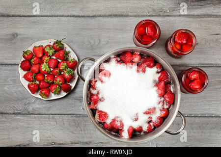 Vue sur table, grand pot de fraises en acier recouvert de cristaux de sucre, trois flacons avec fruits au sirop et la plaque à côté. La fraise fait maison Banque D'Images