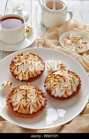 Close-up de mini tartes au citron meringuée doré avec peaks servi avec du thé, mini guimauves et brown sucre biologique sur table en bois avec lumière Banque D'Images