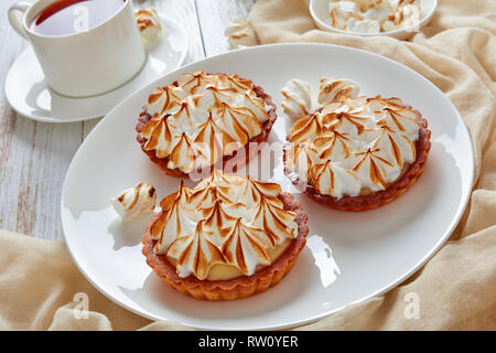Close-up de meringue mini tartelettes dorées avec des pics de meringue servi avec du thé, mini guimauves et brown sucre biologique sur table en bois avec lumière beig Banque D'Images