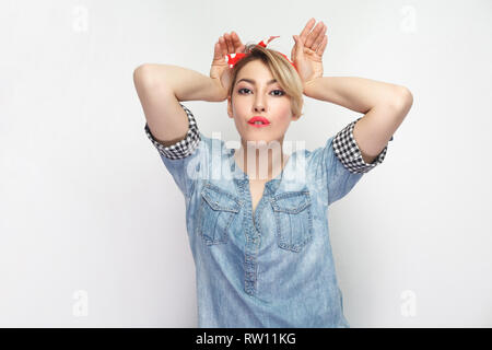 Portrait de funny belle jeune femme en chemise en denim bleu décontracté avec un miroir et bandeau rouge et permanent à la caméra au geste avec oreilles de lapin Banque D'Images