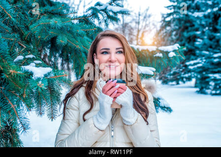 Heureux et smiling girl blouson d'hiver avec les cheveux longs. Dans ses mains tenant une tasse de café de thé. Toile de fond contre les arbres de Noël vert enneigés Banque D'Images