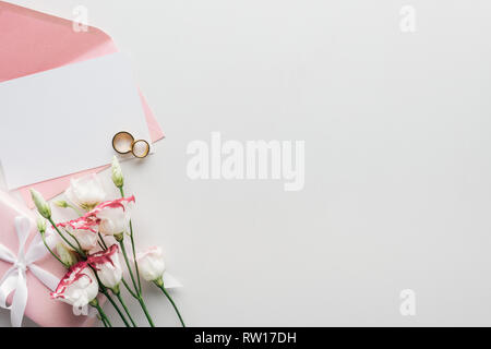 Vue de dessus de la carte vide avec enveloppe rose, fleurs, cadeaux enveloppés et anneaux de mariage d'or sur fond gris Banque D'Images