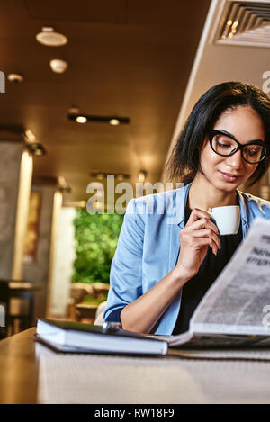 Portrait du jeune chef élégant veste bleu en souriant, tout en regardant à travers des articles dans un journal. Elle est assise à table cafe, boire de morni Banque D'Images