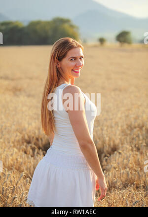 Jeune femme en robe blanche à l'arrière par-dessus son épaule, souriant, le soleil l'après-midi allumé champ de blé derrière elle. Banque D'Images