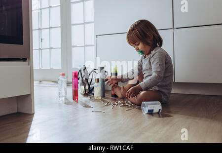 Kid sitting on floor à la maison à jouer avec les oreillettes et les cosmétiques. Enfant jouant avec des articles de ménage assis seul à la maison. Banque D'Images