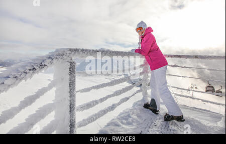 Jeune femme en rose veste de ski, bottes gants et lunettes hat leaning on fence couvertes de neige de cristal de fer, à l'arrière. Soleil qui brille au-dessus de la montagne Banque D'Images