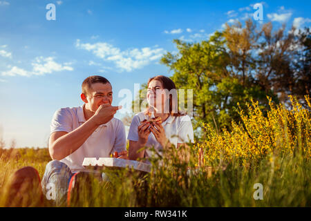 Jeune couple eating pizza et discuter à l'extérieur. La femme et l'homme having picnic au coucher du soleil. Les gars se détendre et profiter de la nourriture. Vie Banque D'Images