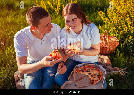 Jeune couple eating pizza et discuter à l'extérieur. La femme et l'homme having picnic au coucher du soleil. Les gars se détendre et profiter de la nourriture. Vie Banque D'Images