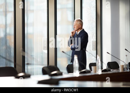 Vue de côté portrait de pensive senior man standing by window in office éclairée par la lumière du soleil, copy space Banque D'Images
