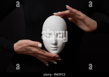 Deux mains tiennent la femme masque de gypse blanc visage sur un fond noir. Masques psychologiques sociaux Concept Banque D'Images