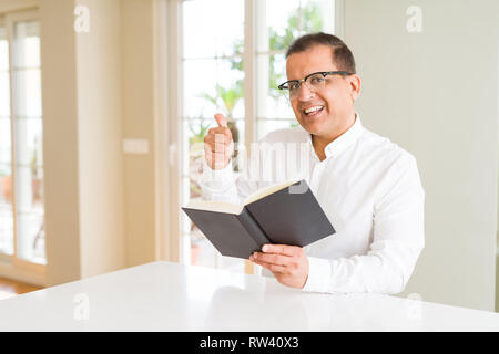 Homme d'âge moyen de la lecture d'un livre à la maison portant des lunettes et de pointage montrant avec le pouce jusqu'à la côte avec happy face smiling Banque D'Images