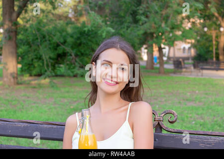 Belle jeune femme est assise sur le banc dans le parc et tenant une bouteille de jus de fruits frais avec une paille. Banque D'Images