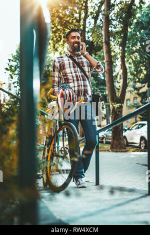 Un beau jeune homme va à la ville avec son vélo, marcher à côté d'elle et de parler sur le smartphone. Banque D'Images