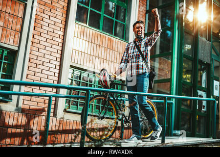Un beau jeune homme va à la ville avec son vélo, debout à côté d'elle et célèbre son succès. Banque D'Images