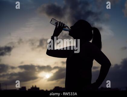Vue de côté de silhouette de jeune femme en vêtements de l'eau potable de la bouteille et merveilleux ciel avec nuages en soirée Banque D'Images