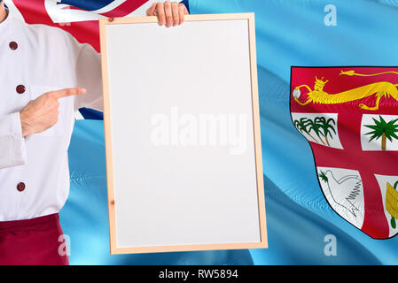 Le fidjien Chef holding blank menu tableau blanc sur fond du drapeau Fidji. Faire cuire en uniforme faisant de l'espace pour le texte. Banque D'Images