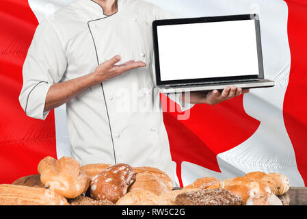 Canadian Baker holding laptop sur drapeau du Canada et de pains. Chef en uniforme faisant écran vide pour copier l'espace. Banque D'Images