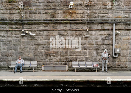 Bien habillée homme plus âgé lire son journal tandis qu'un jeune homme est assis à proximité en utilisant son téléphone intelligent sur la plate-forme de la gare de Charing Cross à Glasgow. Banque D'Images