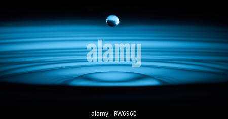 Une seule goutte d'eau gelée dans le temps au milieu de l'air, les ondes à l'eau ci-dessous irradiant vers l'extérieur. Éclairé avec lumière bleue pour donner un look plus abstrait. Banque D'Images