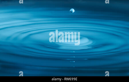 Une seule goutte d'eau gelée dans le temps au milieu de l'air, les ondes à l'eau ci-dessous irradiant vers l'extérieur. Éclairé avec lumière bleue pour donner un look plus abstrait. Banque D'Images
