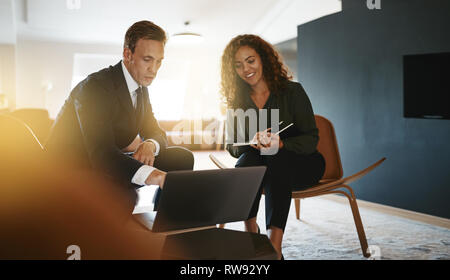 Deux young businesspeople smiling tout en parlant et en travaillant sur un ordinateur portable ensemble dans un bureau moderne et lumineux Banque D'Images