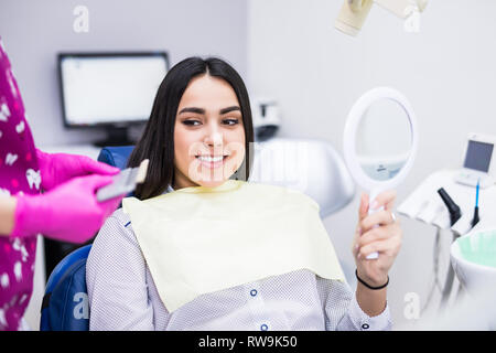 Belle jeune femme avec des dents blanches parfaites assis dans fauteuil dentaire, à la recherche dans le miroir et contrôler les dents après un traitement au modern Banque D'Images