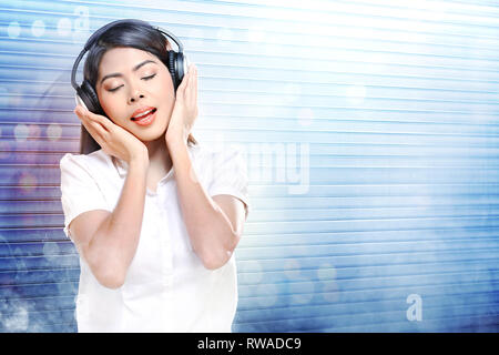 Jolie femme asiatique en chemise blanche à l'écoute de la musique avec des écouteurs sur rouleau blanc porte d'obturation avec de la fumée et de réflexion de la lumière colorée background Banque D'Images