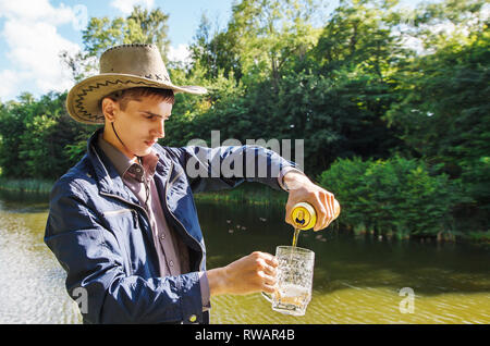 Jeune homme dans un chapeau de cowboy est servir la bière d'un jar dans un mug dans le parc aux beaux jours d'été Banque D'Images