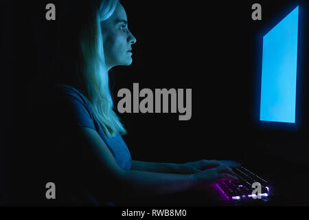 Blonde femme assise en face de moniteur du PC, les mains sur le clavier, travailler, illuminé par la lumière de l'écran bleu Banque D'Images