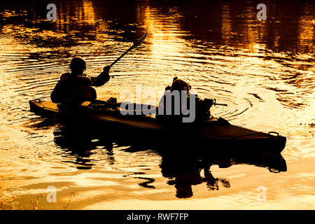 Silhouette d'hommes des forces spéciales de l'Armée de pagaie kayak sur la rivière au crépuscule. Banque D'Images