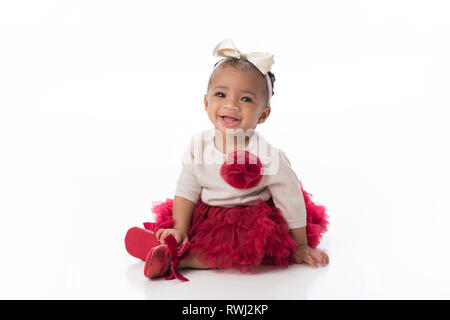 A smiling six mois baby girl wearing a red tutu. Elle est assise sur un arrière-plan transparent, blanc. Banque D'Images