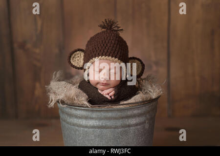 Une semaine bébé nouveau-né garçon portant un chapeau brun, singe en crochet. Il est en train de dormir dans un seau en acier galvanisé et a un léger sourire sur son visage. Banque D'Images