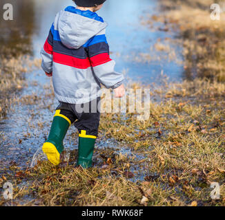 Petit garçon marcher dans des flaques dans l'herbe portant des bottes de pluie. Banque D'Images