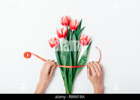 Lier les mains de la femme bouquet de fleurs tulipes rouges frais avec le satiné sur table en bois blanc, vue du dessus. Banque D'Images