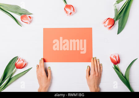 Mise à plat de la composition de la femme avec les mains manucure vert feuille de papier rouge holding blank près de magnifiques tulipes sur table en bois blanc, vue du dessus. Banque D'Images