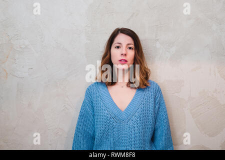 Jolie jeune femme sérieuse en chandail tricoté à la caméra au mur gris et, debout près de Banque D'Images