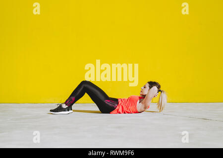 Jeune femme dans les vêtements de sport faisant asseoir ups exercices sur terre près de mur jaune Banque D'Images