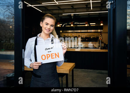 Serveuse Portant tablier, debout à l'entrée du restaurant. Femme assister à de nouveaux clients dans son café. Happy woman showing open sign dans sa petite entreprise Banque D'Images