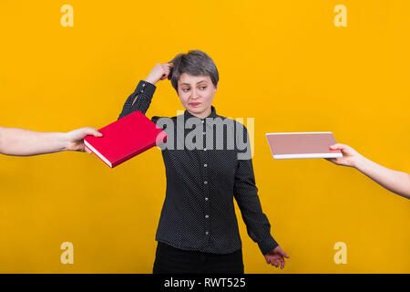 Pensive young woman student doigt à la tête de penser à ce que de choisir entre un livre et l'ordinateur tablette plus isolé mur jaune. Savoirs traditionnels Banque D'Images