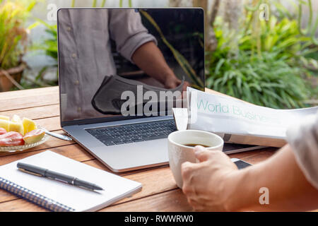 Businessman hand holding coffee lisant le journal avec un ordinateur portable sur la table. Banque D'Images