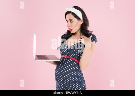 Surpris femme enceinte en notation dress looking at laptop écran isolé sur pink Banque D'Images