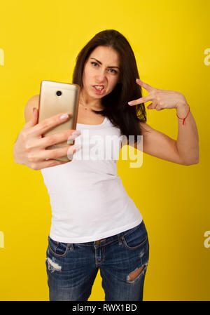 Charmante jeune fille brune dans des tenues à la mode fait une sur le téléphone et selfies grimaces sur un fond jaune lumineux Banque D'Images